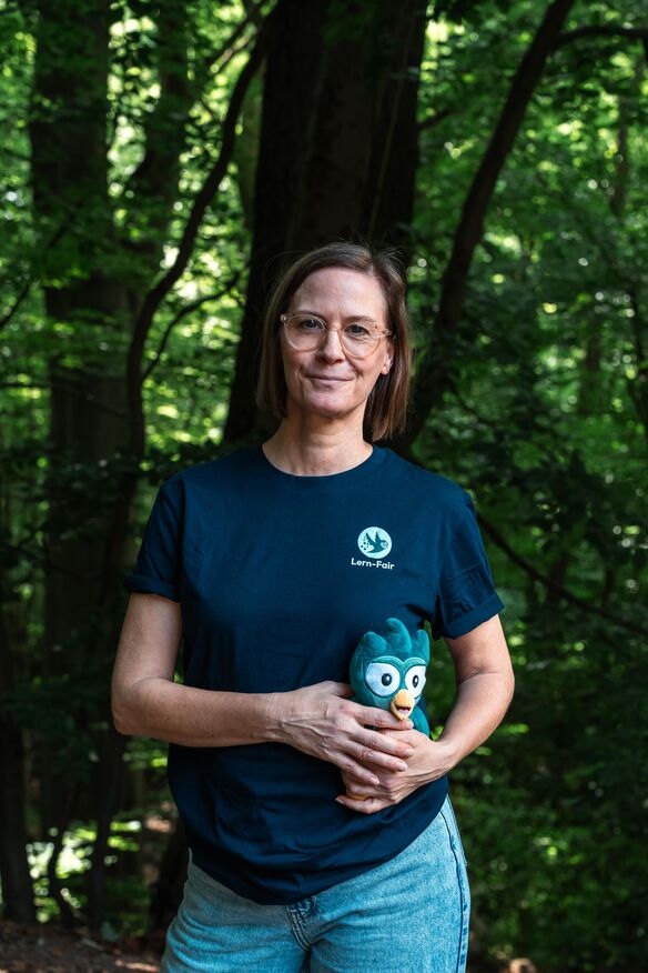 Junge Frau in Lern-Fair T-Shirt vor Wald mit Eulen Maskottchen
