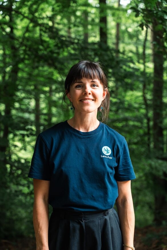 Junge Frau in Lern-Fair T-Shirt vor Wald mit Eulen Maskottchen