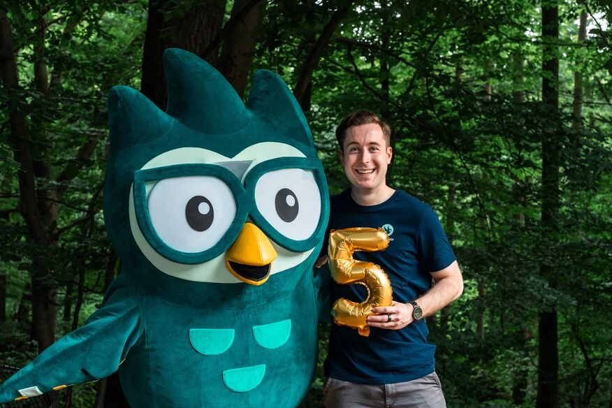 Junger Mann in Lern-Fair T-Shirt vor Wald mit Eulen Maskottchen