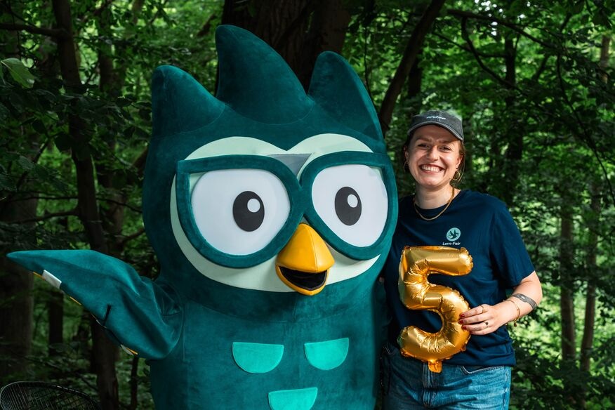 Junge Frau in Lern-Fair T-Shirt vor Wald mit Eulen Maskottchen