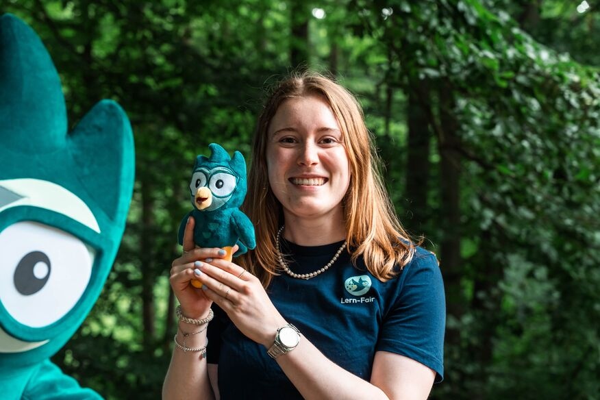 Junge Frau in Lern-Fair T-Shirt vor Wald mit Eulen Maskottchen