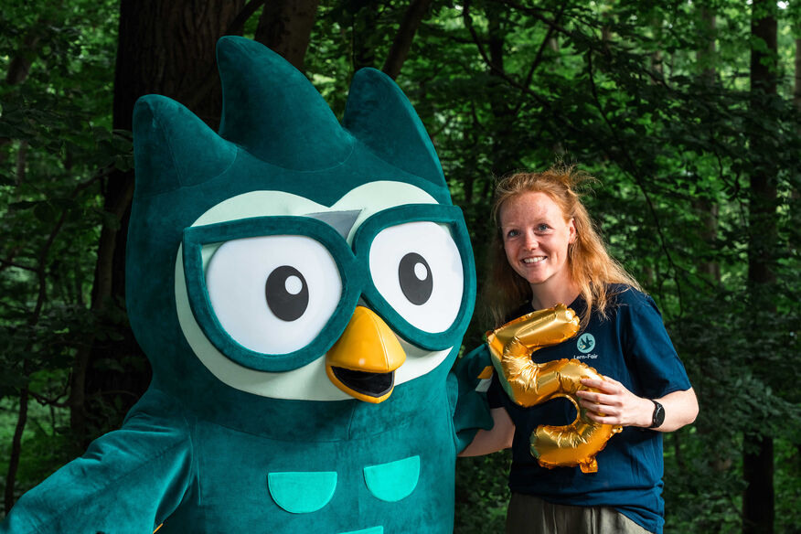 Junge Frau in Lern-Fair T-Shirt vor Wald mit Eulen Maskottchen