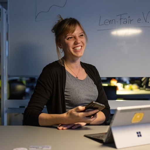 Junge Frau mit mobilem Endgerät in der Hand lächelt in die Kamera vor Schriftzug Lern-Fair.