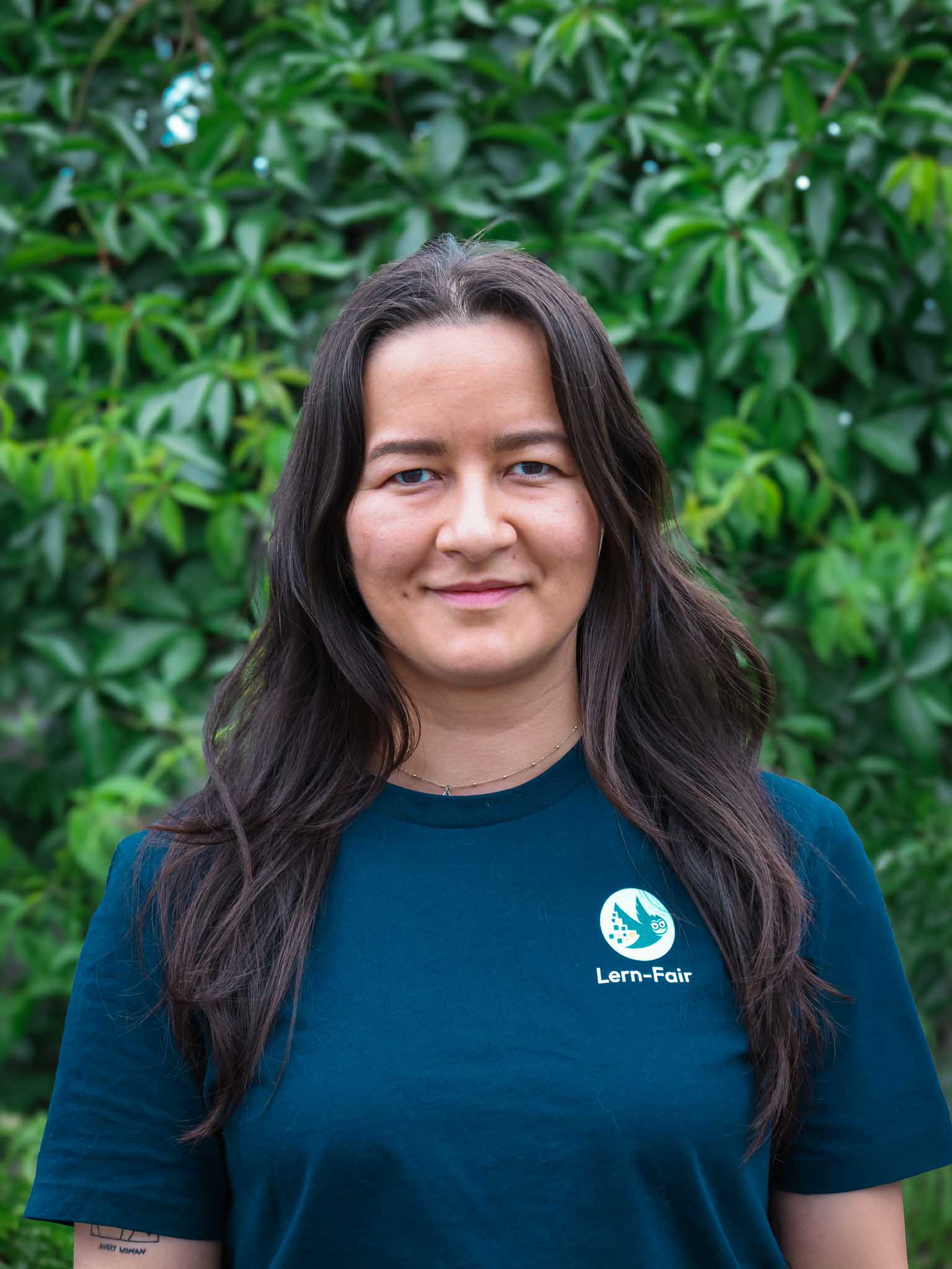 Junge Frau in Lern-Fair T-Shirt  mit Eule als Logo steht vor grünem Hintergrund aus Pflanzen.