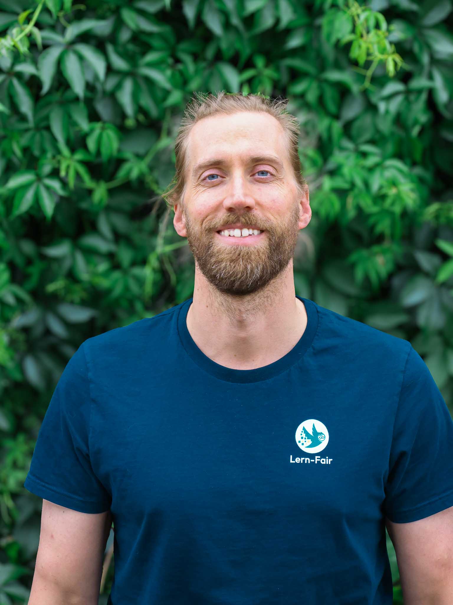 Junger Mann in Lern-Fair T-Shirt  mit Eule als Logo steht vor grünem Hintergrund aus Pflanzen.