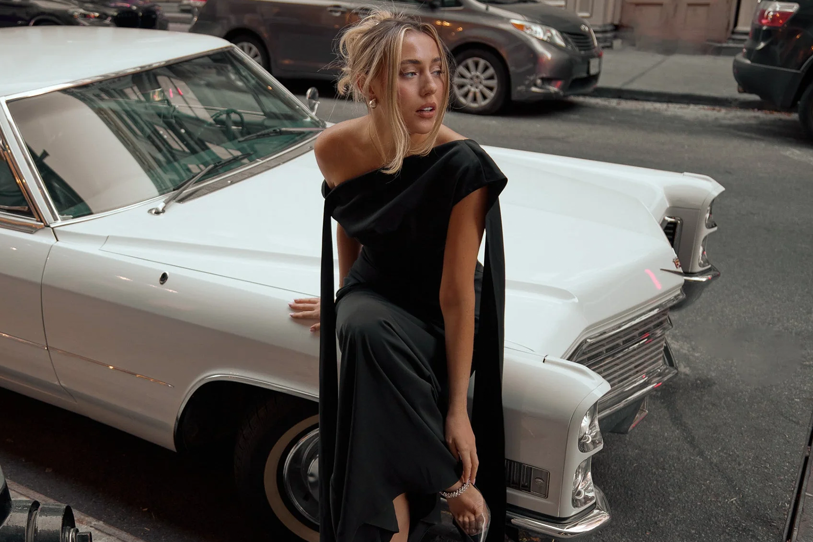 Model in a black off-the-shoulder Odd Muse dress leaning against a white vintage car