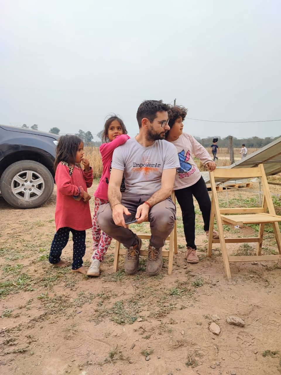 Hombre sentado en una silla con tres niñas alrededor en un campo árido al aire libre.
