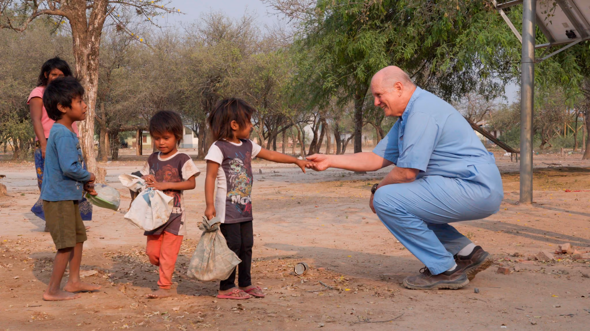 Hombre sonriente en ropa médica saluda a niña mientras otros niños observan al aire libre.