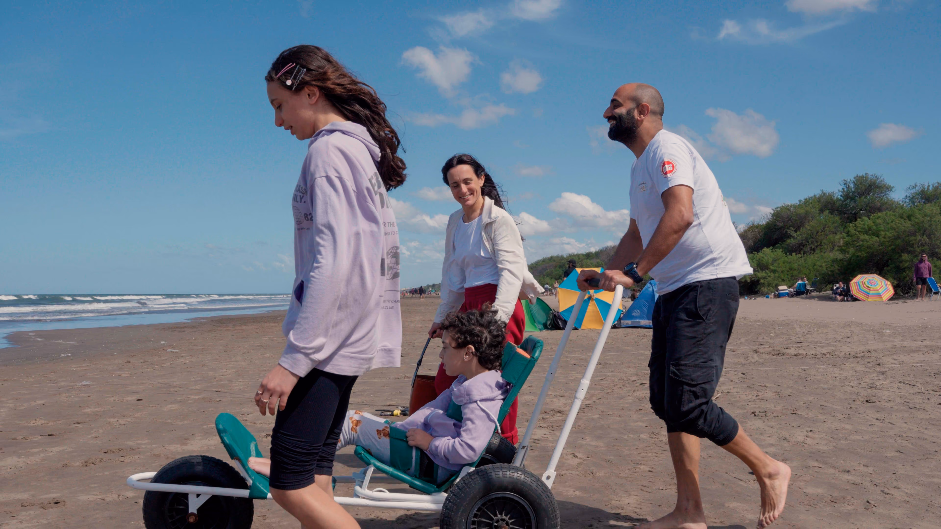 Familia feliz en la playa con hombre empujando carro, niños y mujer caminando en arena bajo cielo azul