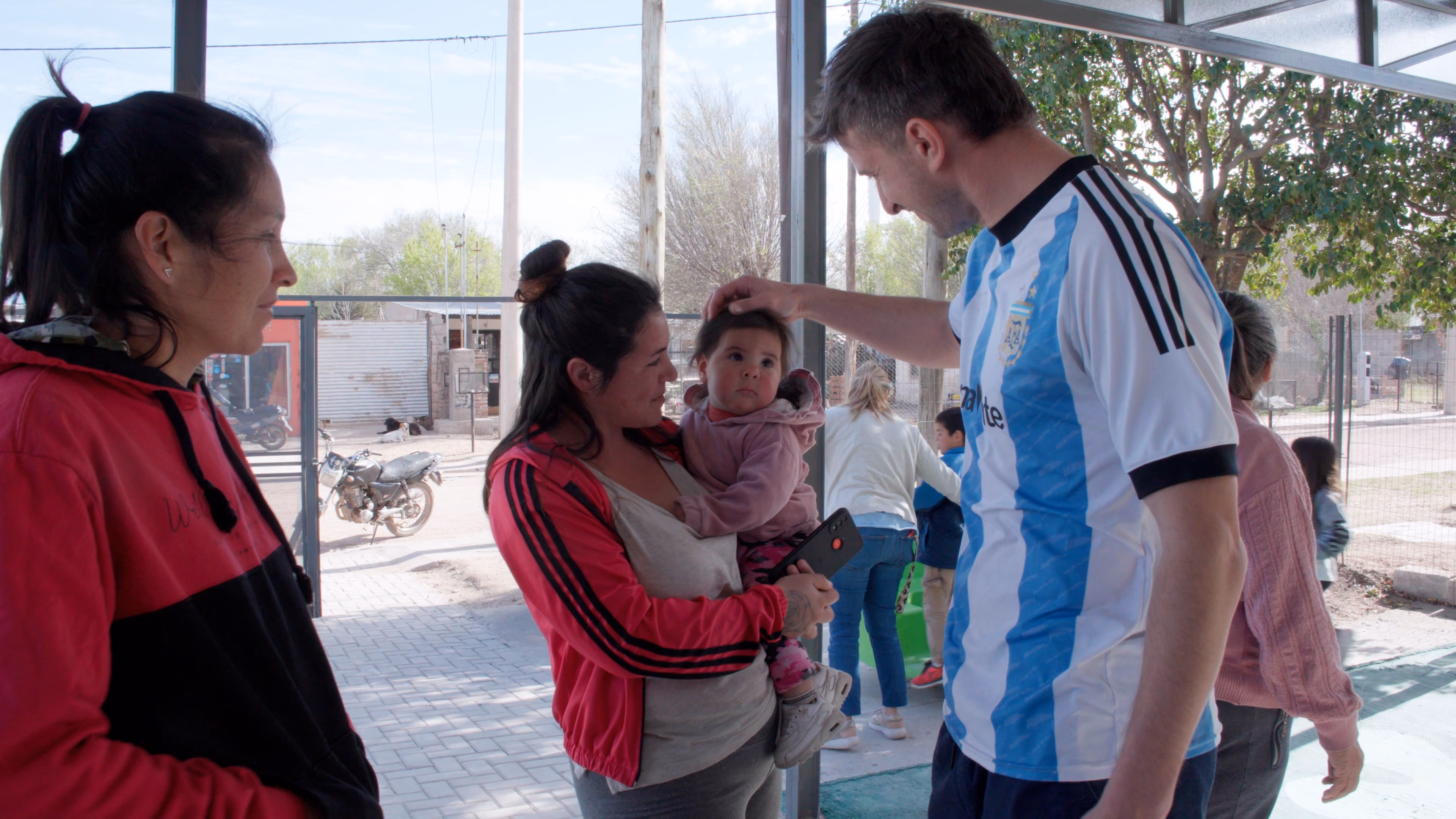 Un hombre con camiseta de Argentina acaricia la cabeza de un bebé que sostiene una mujer con chaqueta roja.