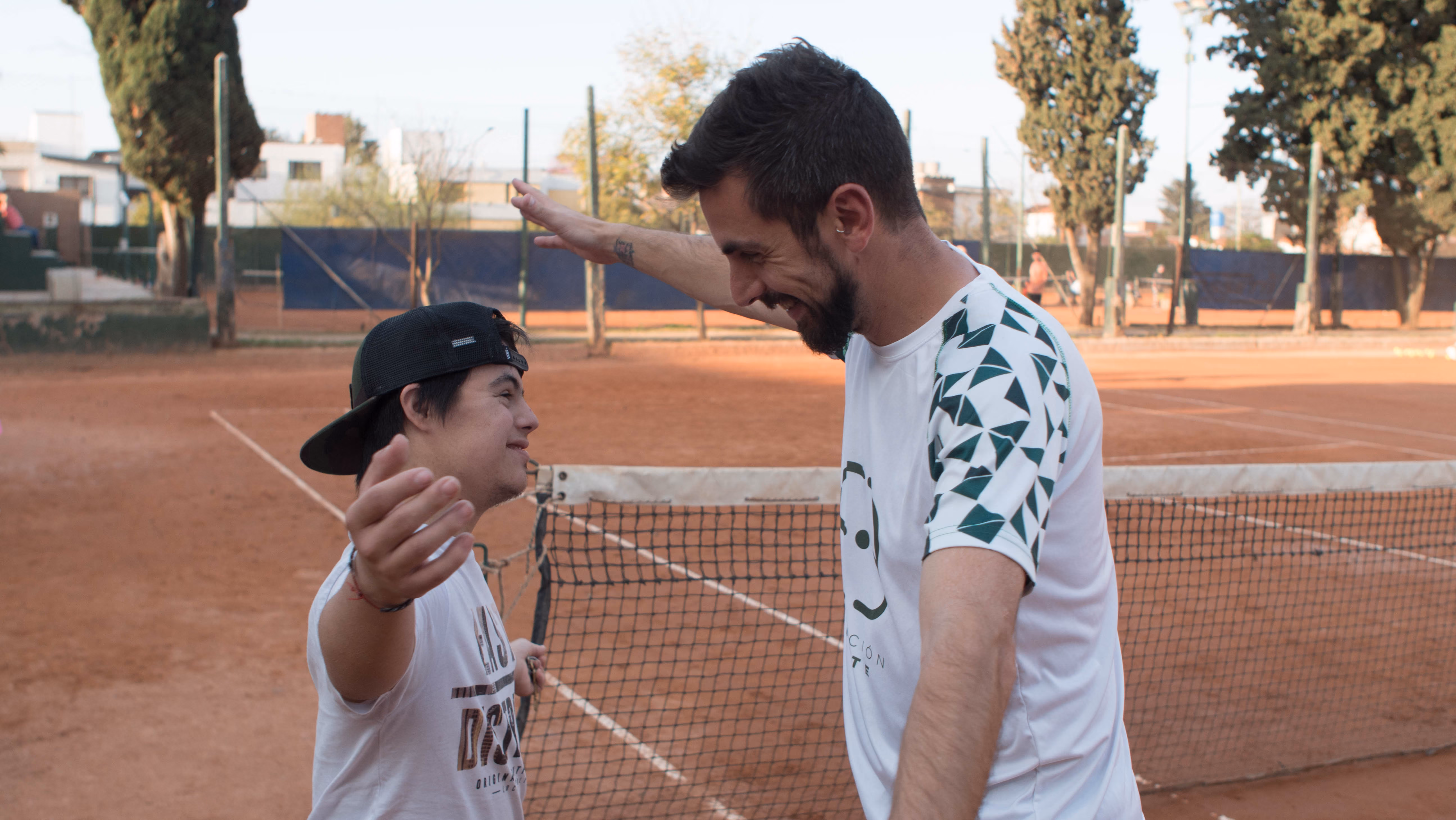 Dos hombres sonrientes celebran con los brazos abiertos en una cancha de tenis de tierra batida.