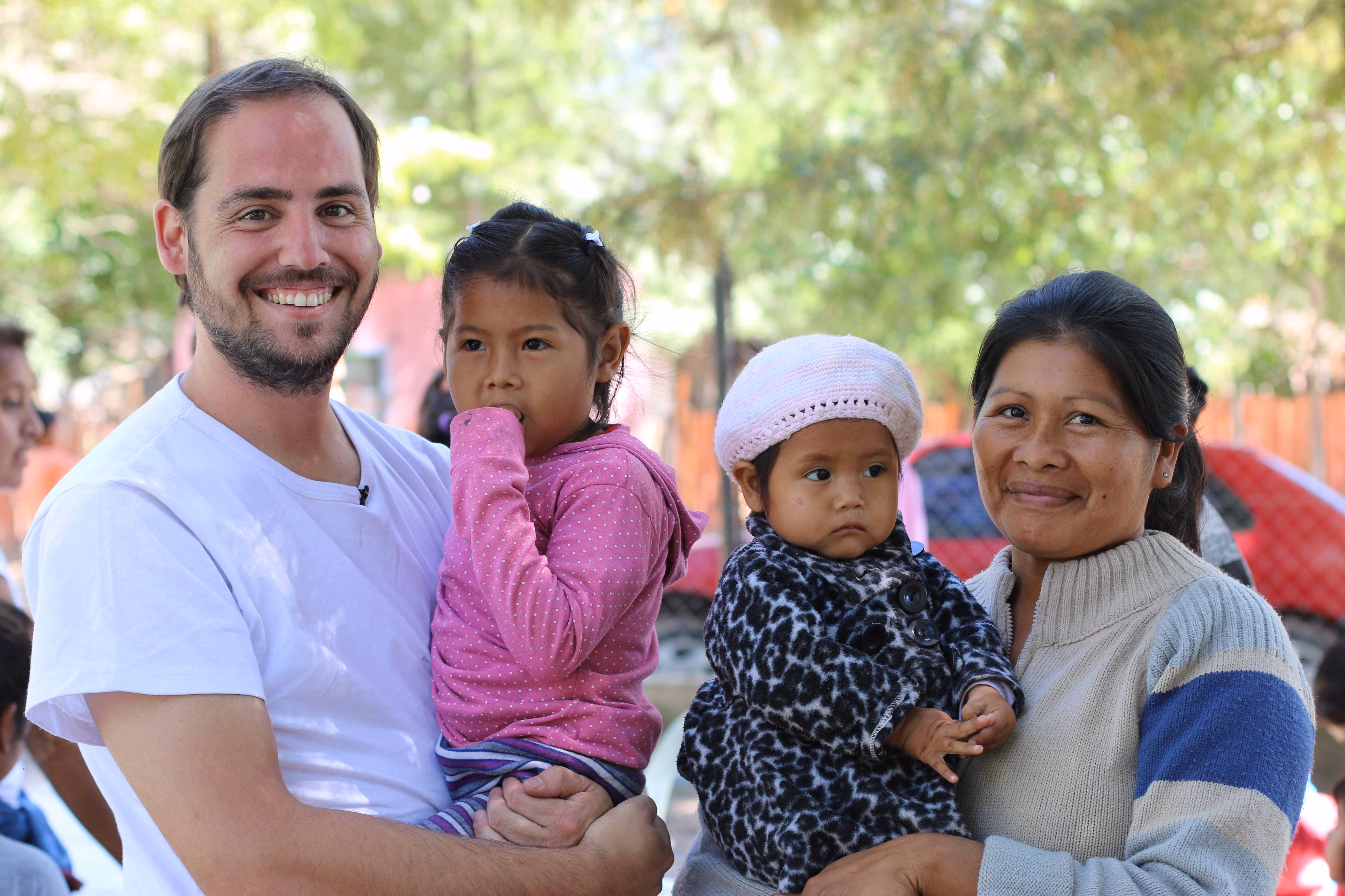 Hombre sonriente y mujer con dos niñas pequeñas en brazos en un parque al aire libre.