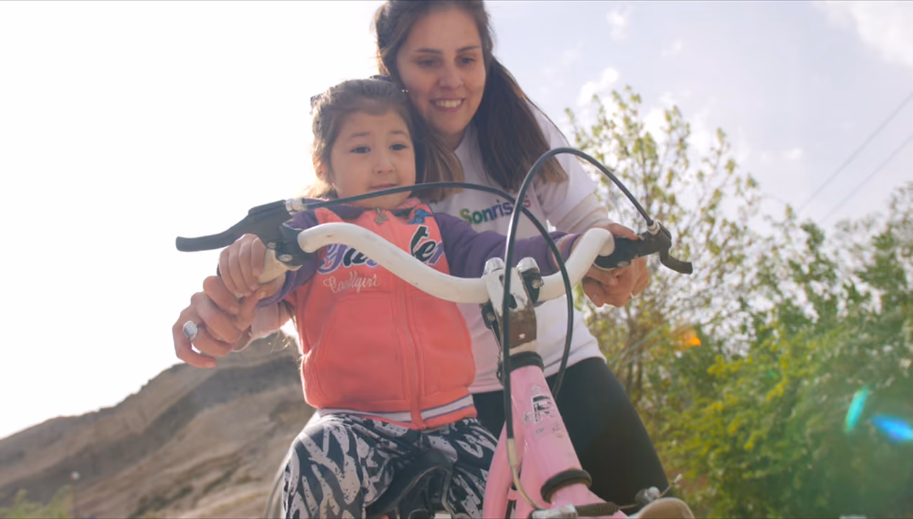 Mujer enseña feliz a niña a andar en bicicleta rosa al aire libre bajo cielo claro.