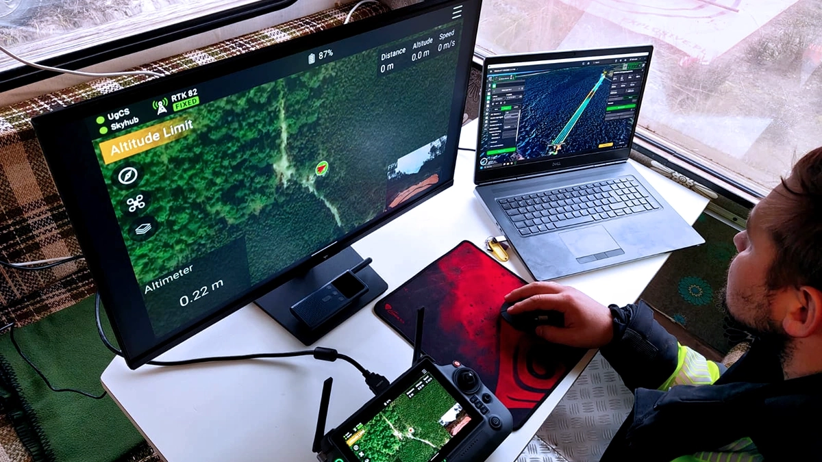 Drone operator monitoring UgCS flight planning software with SkyHub connected and RTK Fixed status during magnetometer UXO survey, DJI M350 visible on landing pad through window