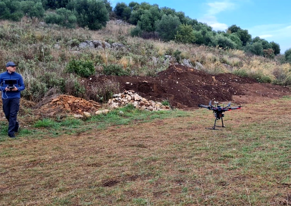 Researcher holding a controller beside a DJI Matrice 600 hexacopter with its LiDAR payload, on cleared ground below the scrub-covered Kastrì-Pandosia hillside
