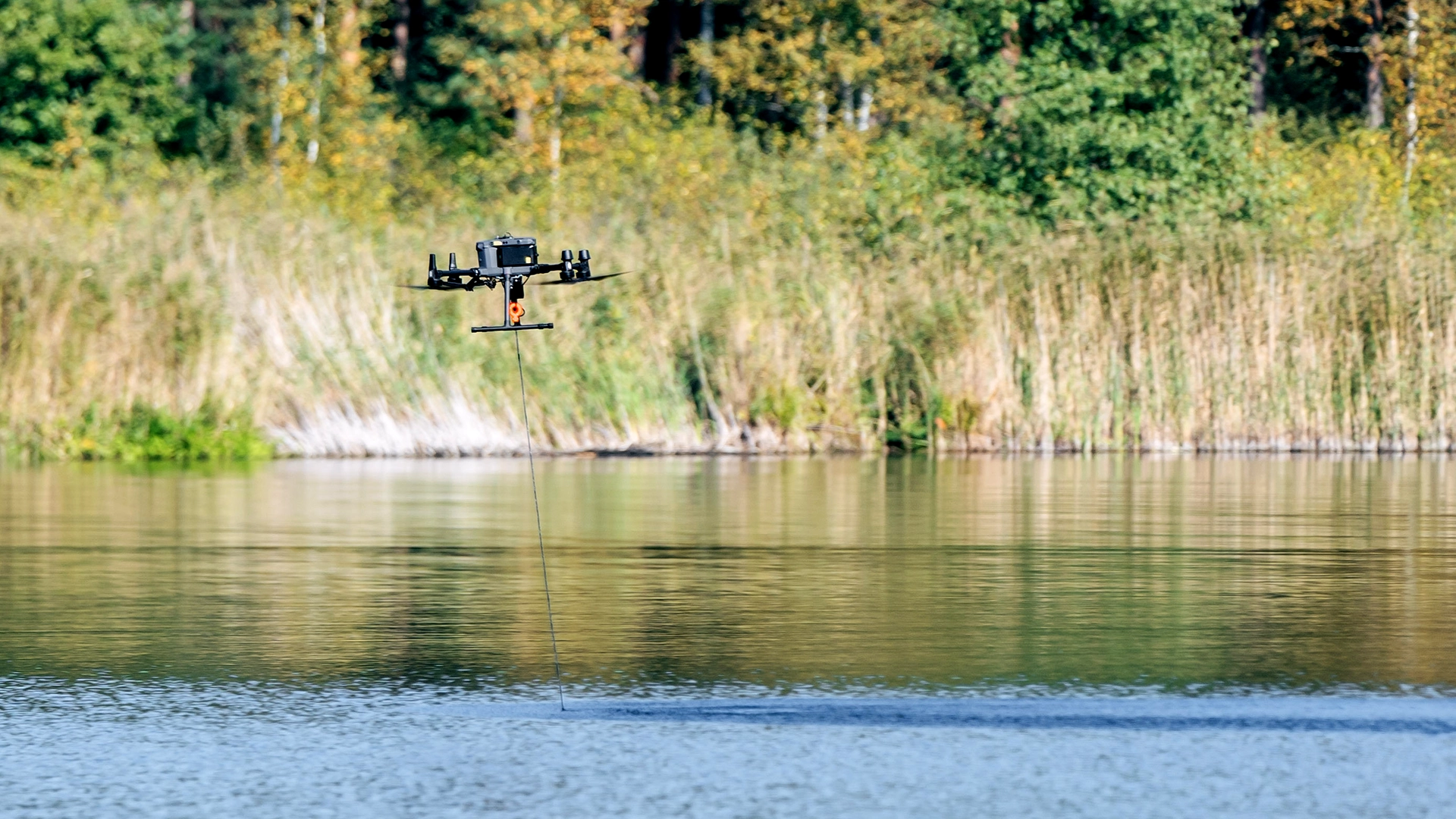 Drone flying low over water with an echo sounder cable running into the water in front of a reed bed