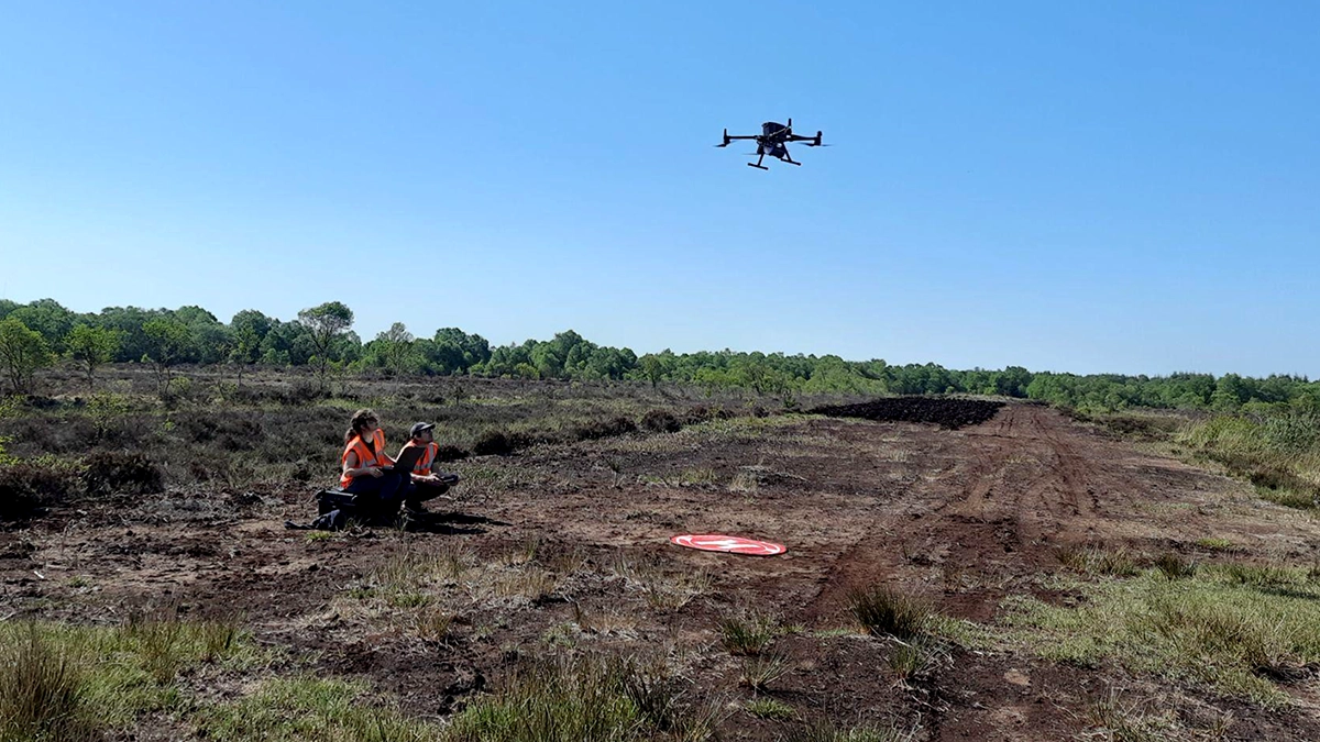 Two survey crew members beside a landing pad with a drone airborne over peatland