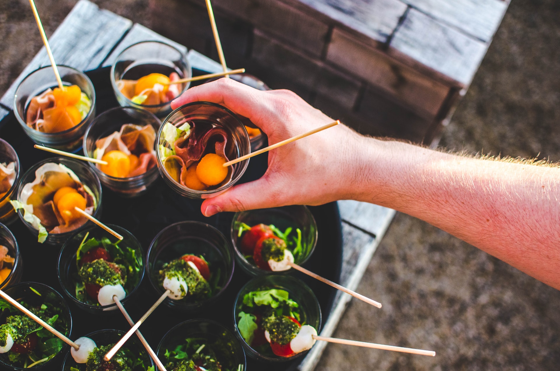 Hand lifting a cup of prosciutto and melon from a tray of appetizer cups