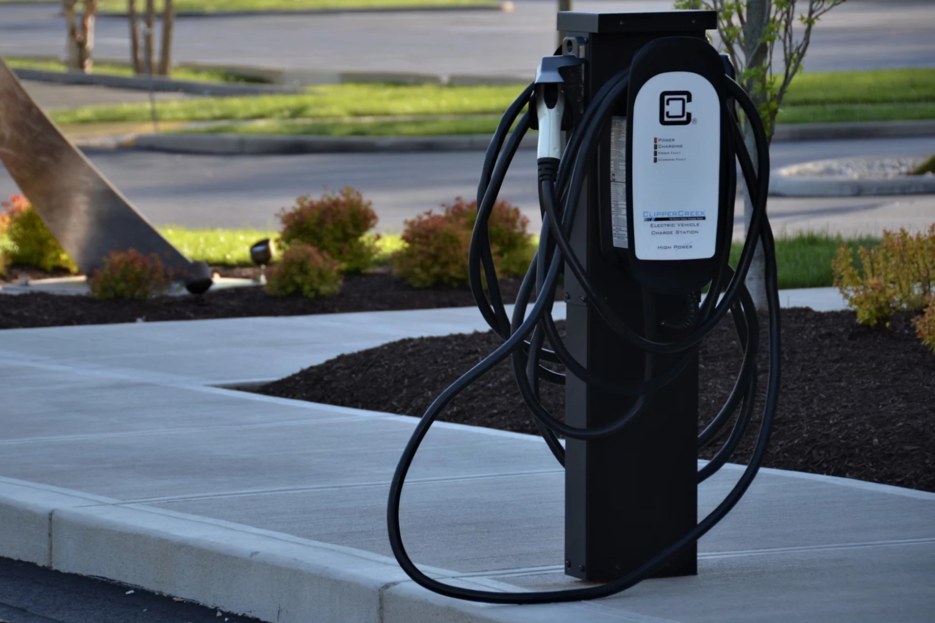 An EV charging unit is located on a sidewalk by a garden.