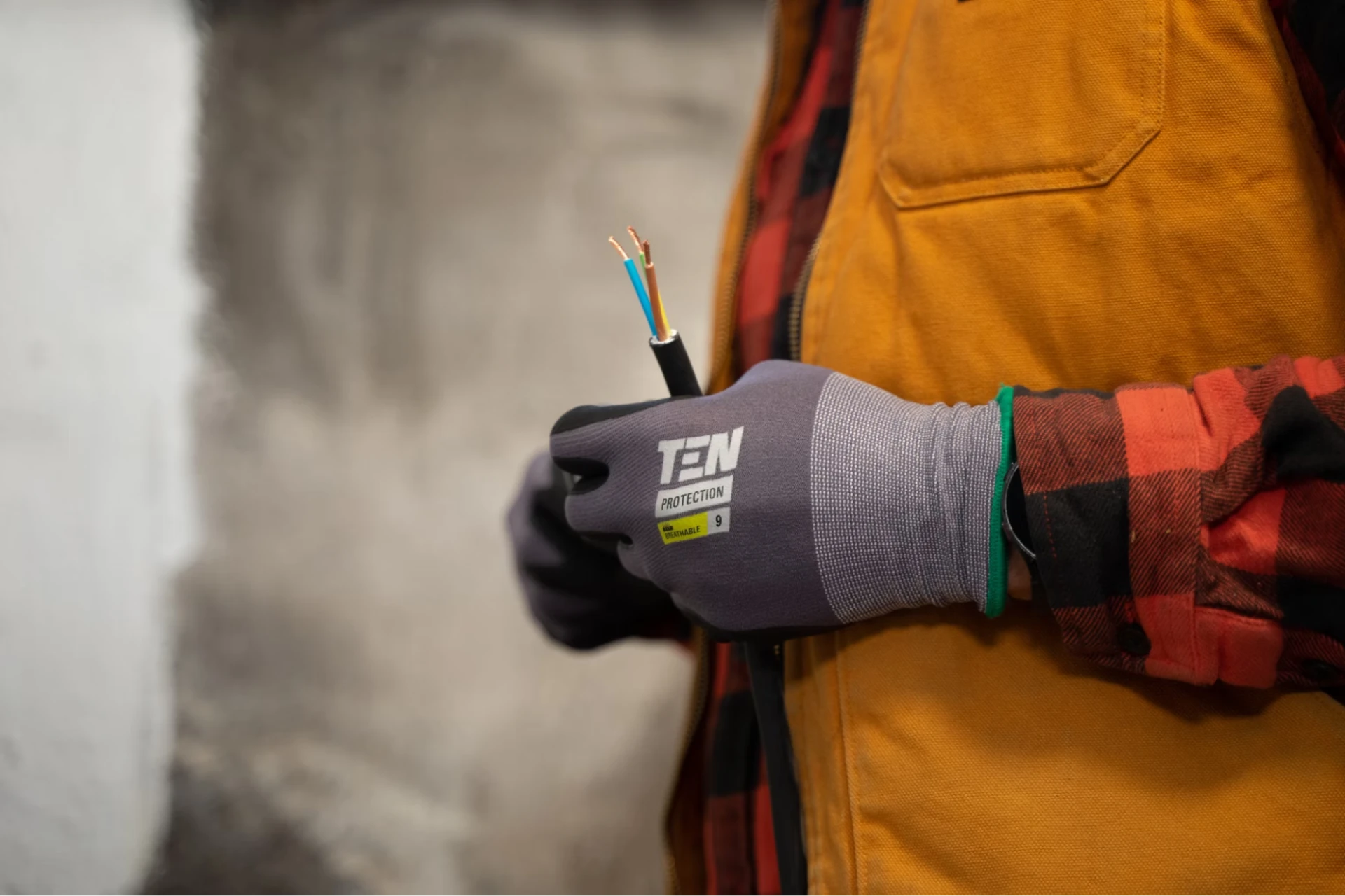 An electrician in beige overalls and orange plaid shirt, holds exposed wires while wearing grey gloves. 