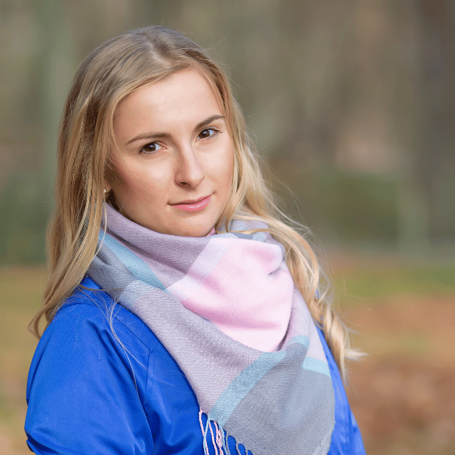 Blonde woman wearing a blue jacket and a pastel pink, gray, and blue scarf outdoors.