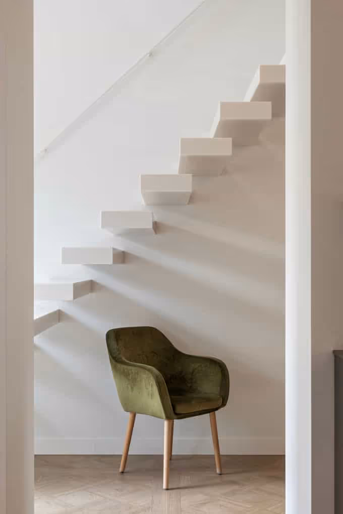 Vastgoedfotografie van een moderne, minimalistische witte trap met een groene fluwelen stoel in de hoek.