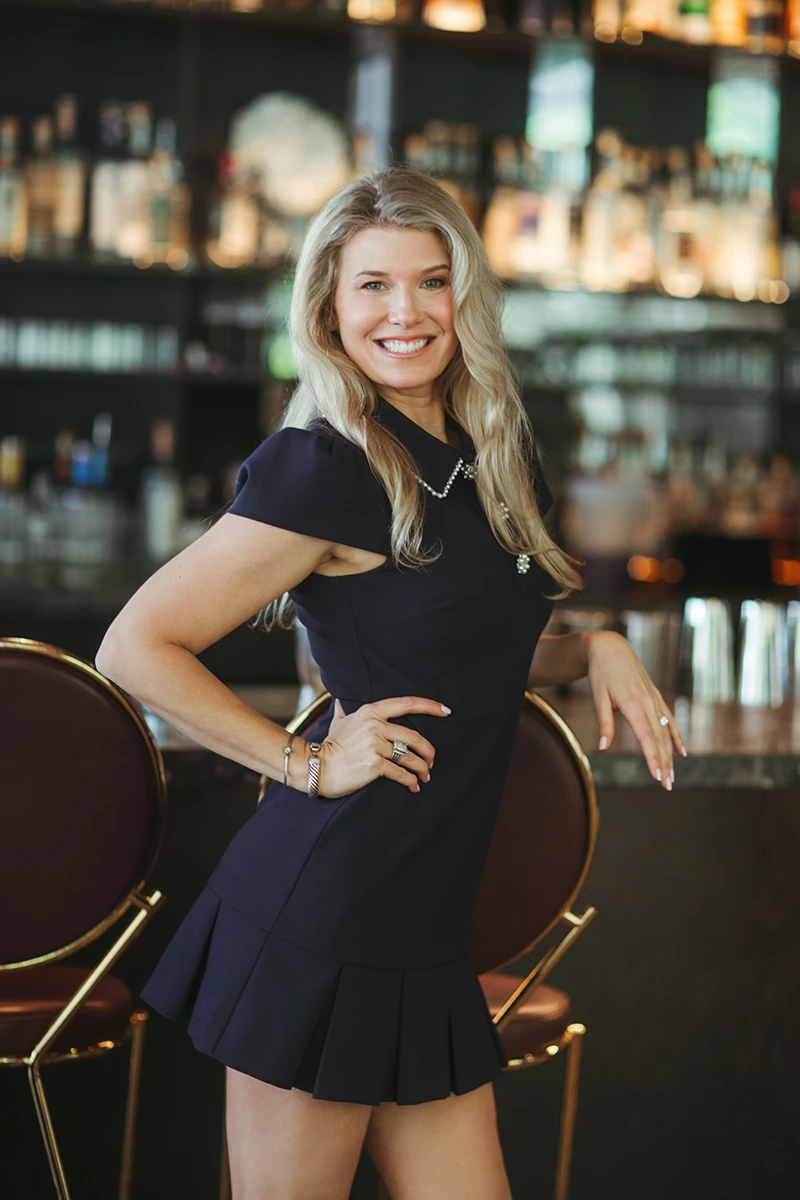 Photo of Ash in a navy dress in front of a beautiful bar.