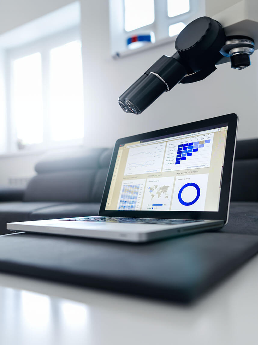 Laptop on a dark surface displaying research data charts with a microscope positioned above it in a bright room.