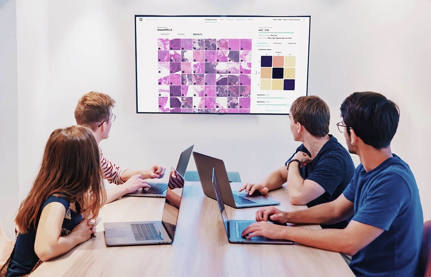 Four people sitting at a conference table with laptops, viewing a large screen displaying histology slide images and data analysis results.