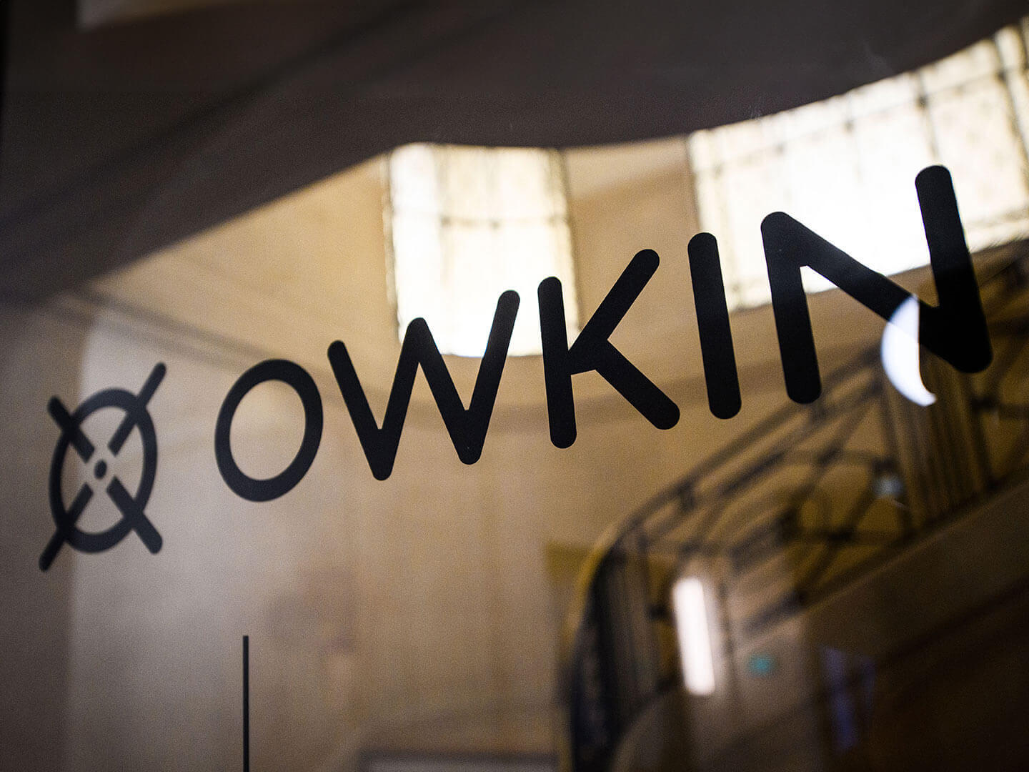 Close-up of a glass panel with the word 'OWKIN' and a circular logo with intersecting lines, with blurred indoor background.