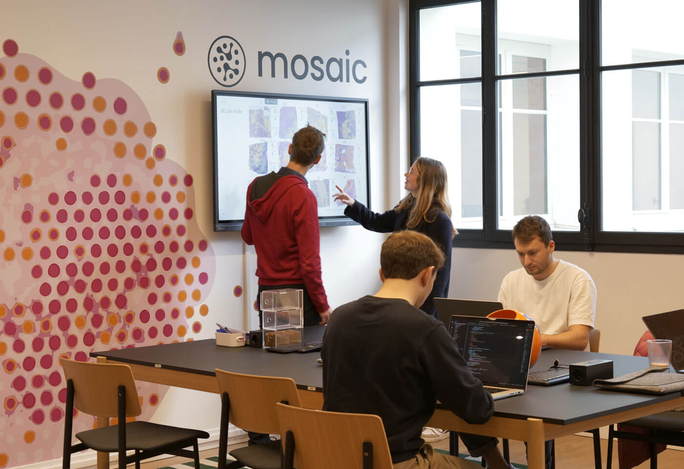 Four people in a modern office; two discuss data on a wall screen near a mosaic logo, while two others work on laptops at a table.