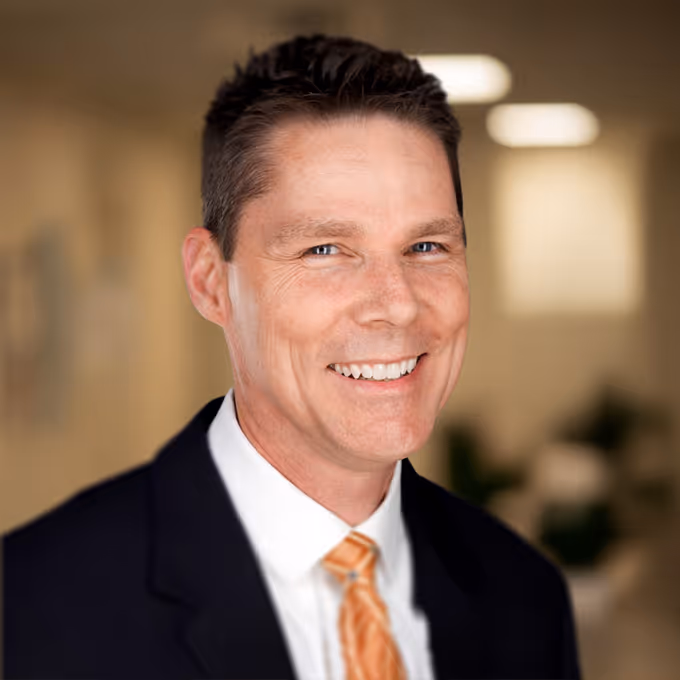 Kerry Morris smiling in a dark suit and orange striped tie with a blurred indoor background.