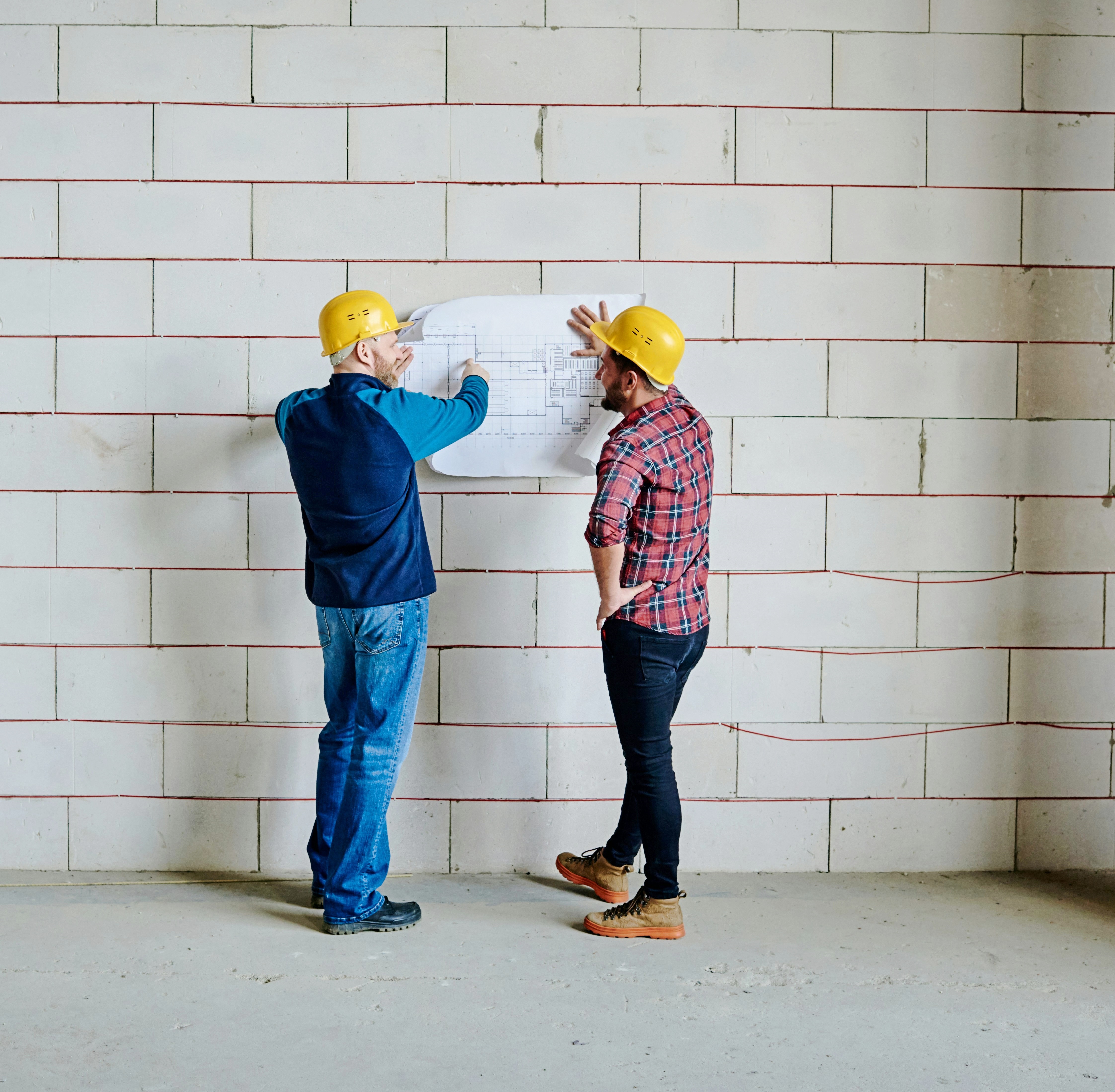 Architekt und Statiker stehen auf der Baustelle mit einem Plan vor einer gemauertem Wand und besprechen sich