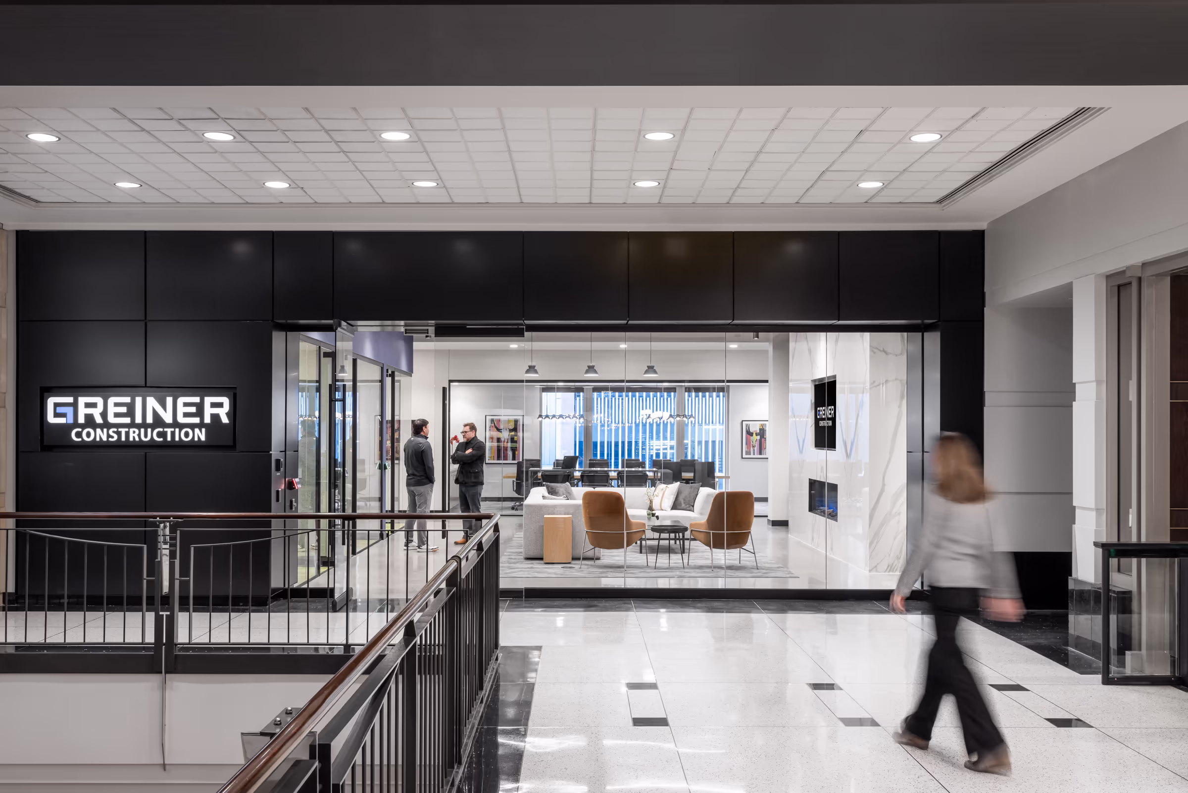 Office entrance with Greiner Construction sign, seating area, two men talking, and a woman walking by.