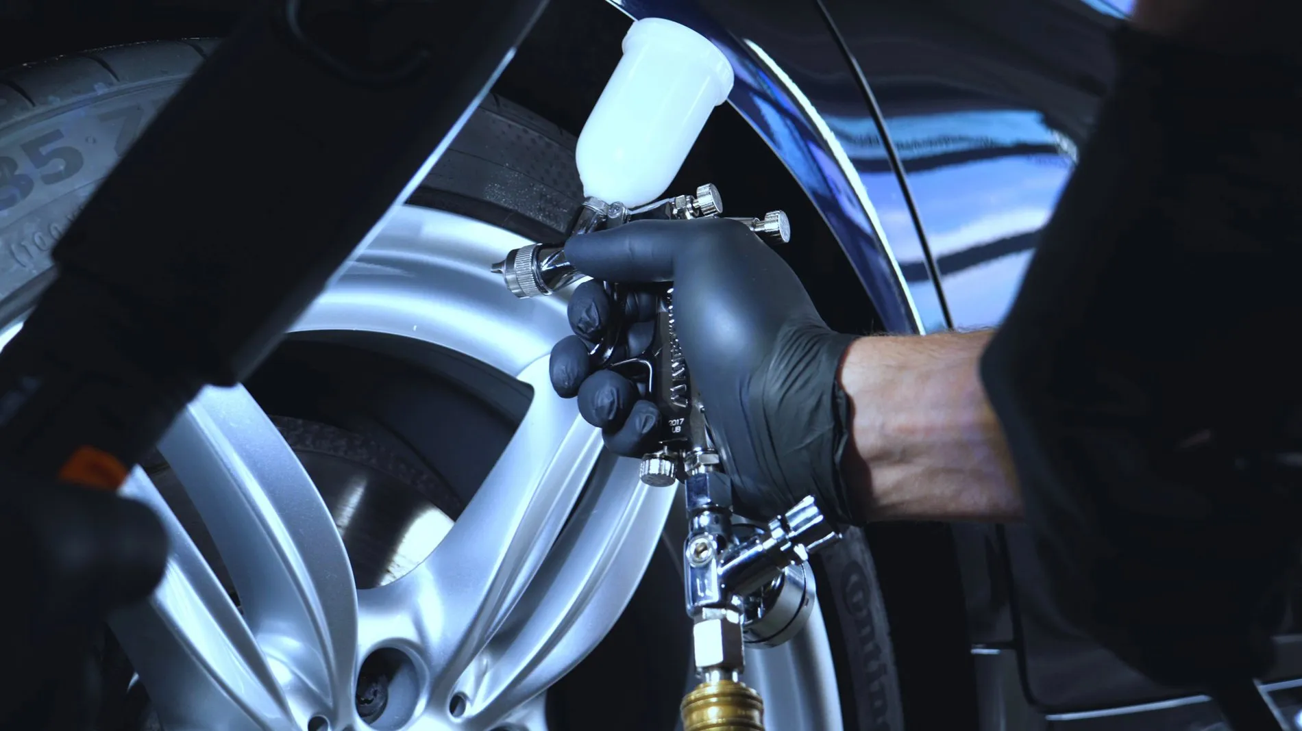 Application d’une peinture ou d’un revêtement sur une jante argentée, avec une main gantée tenant un pistolet pulvérisateur dans un atelier automobile.