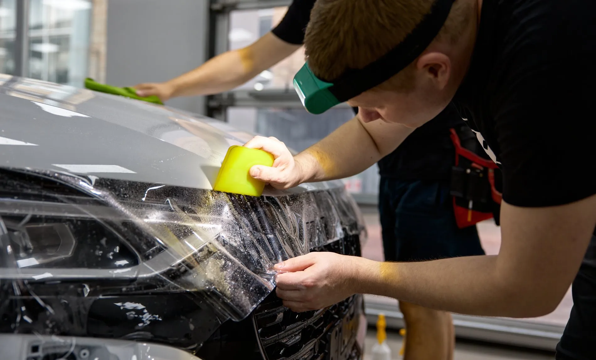 Application d’un film de covering sur le capot d’une voiture blanche par un technicien utilisant une raclette en plastique dans un atelier professionnel.