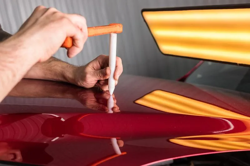 Un professionnel effectuant un débosselage sans peinture sur le capot d'une voiture rouge, utilisant un outil de précision pour restaurer la surface sans abîmer la peinture.