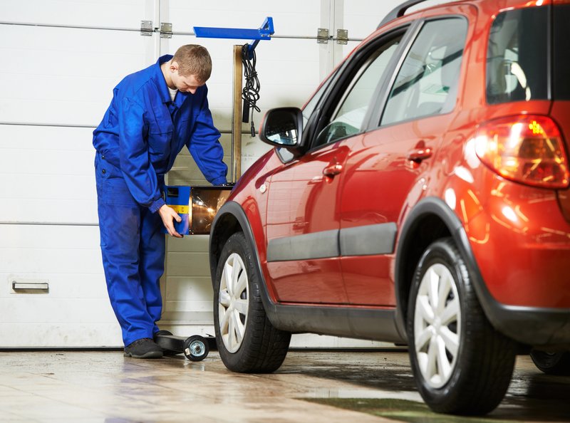 Un technicien en combinaison bleue effectue un contrôle technique des phares d'une voiture rouge dans un garage.