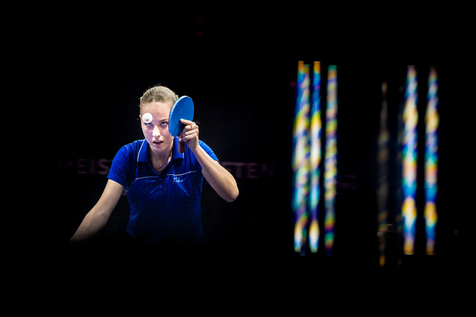 Focused female table tennis player in a blue shirt preparing to serve with paddle and ball near her face against a dark background.