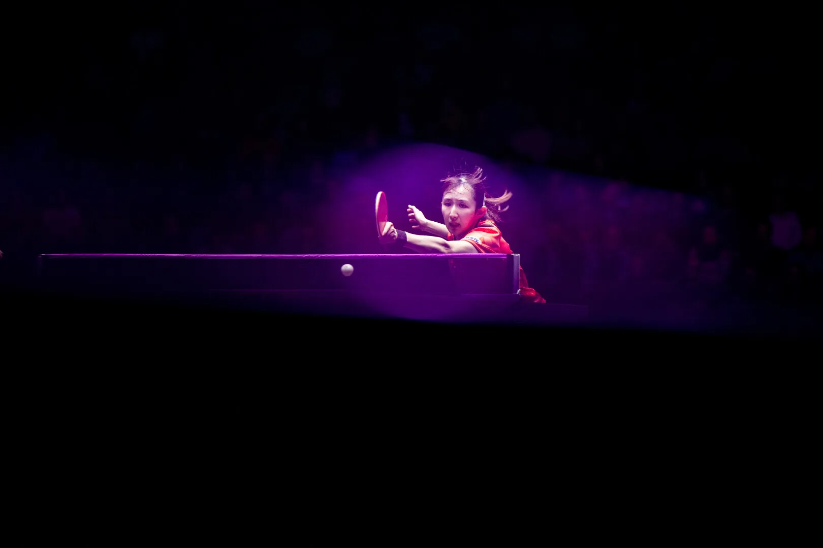 Female table tennis player in red hitting the ball during a match under purple spotlight.