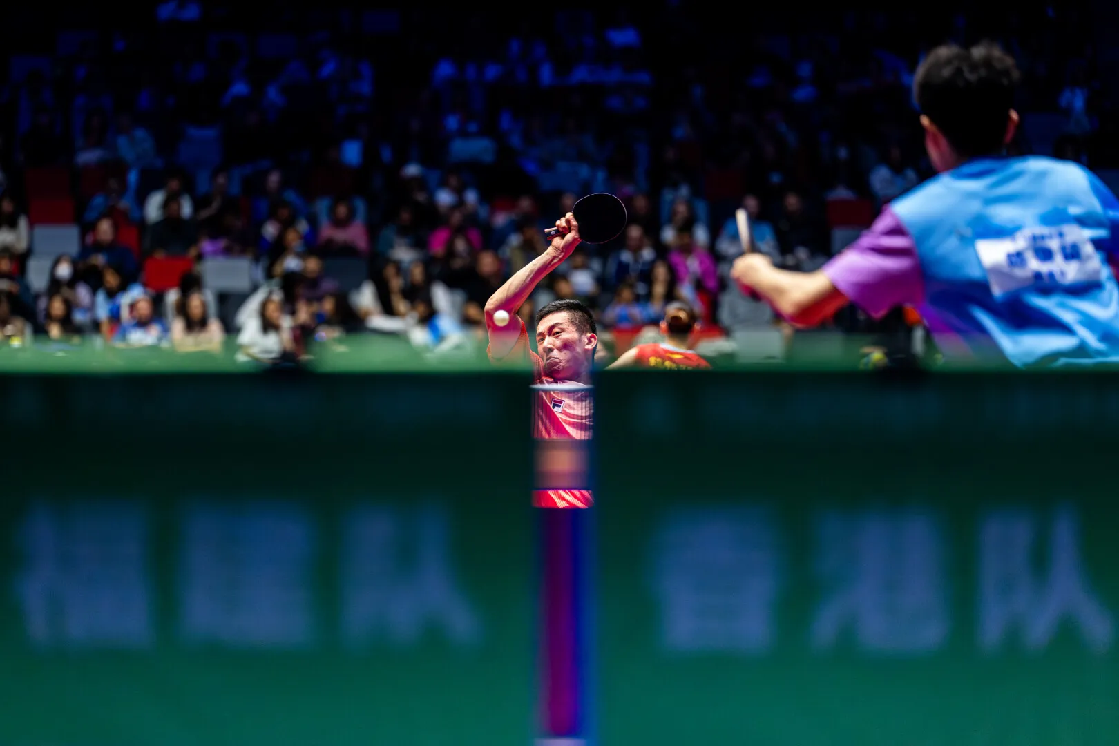 A table tennis player in red prepares to strike the ball mid-air during a competitive match with an audience in the background.