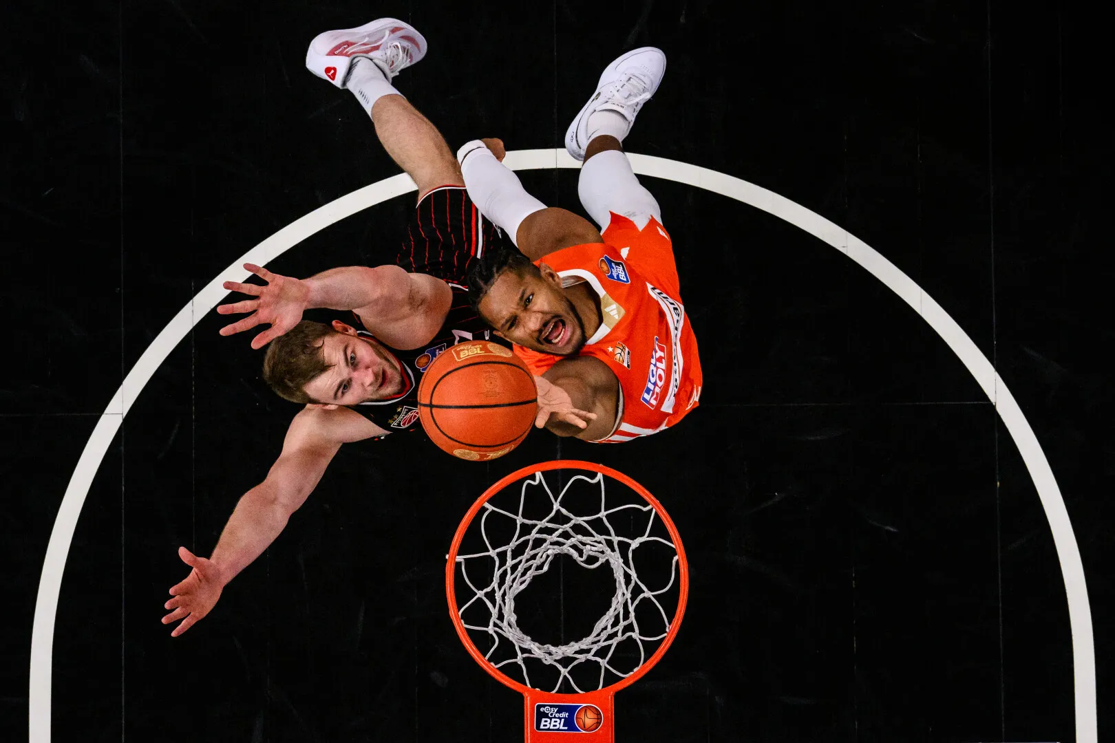 Two basketball players in mid-air reaching for the ball above the hoop during a game.