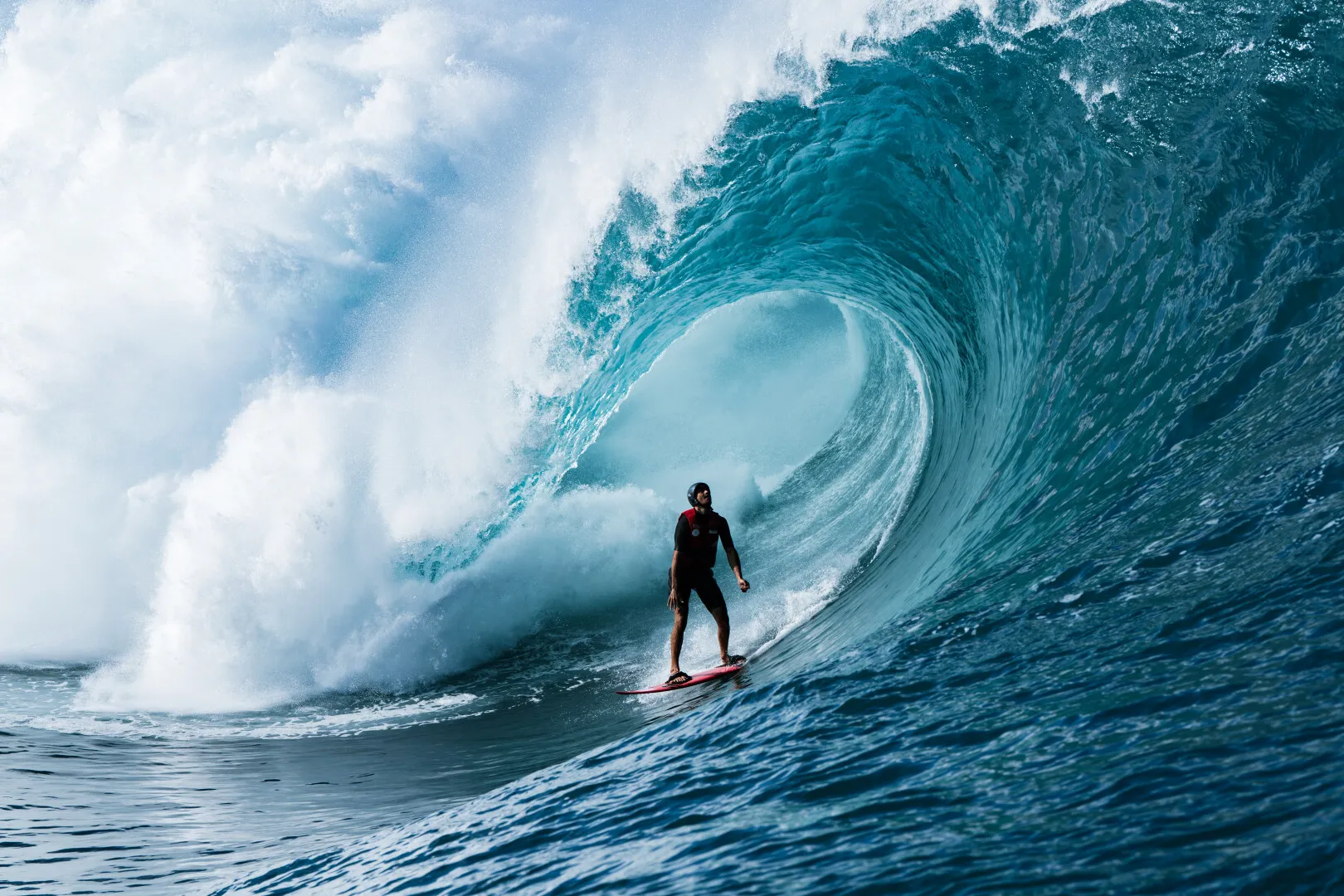 Surfer wearing a helmet riding a large blue ocean wave curled into a tube.