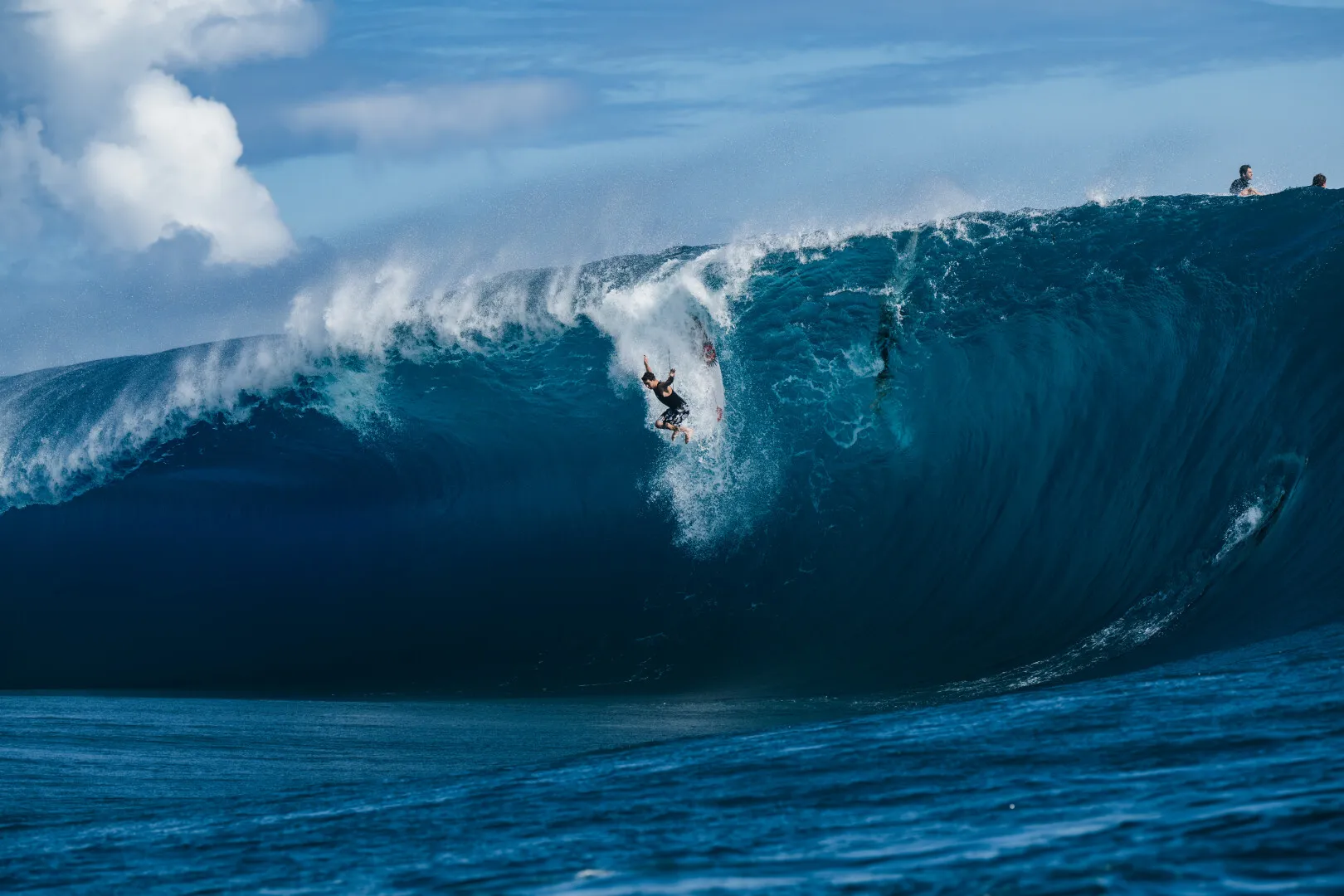 Surfer falling off a large blue wave with two other surfers visible at the wave crest under a partly cloudy sky.