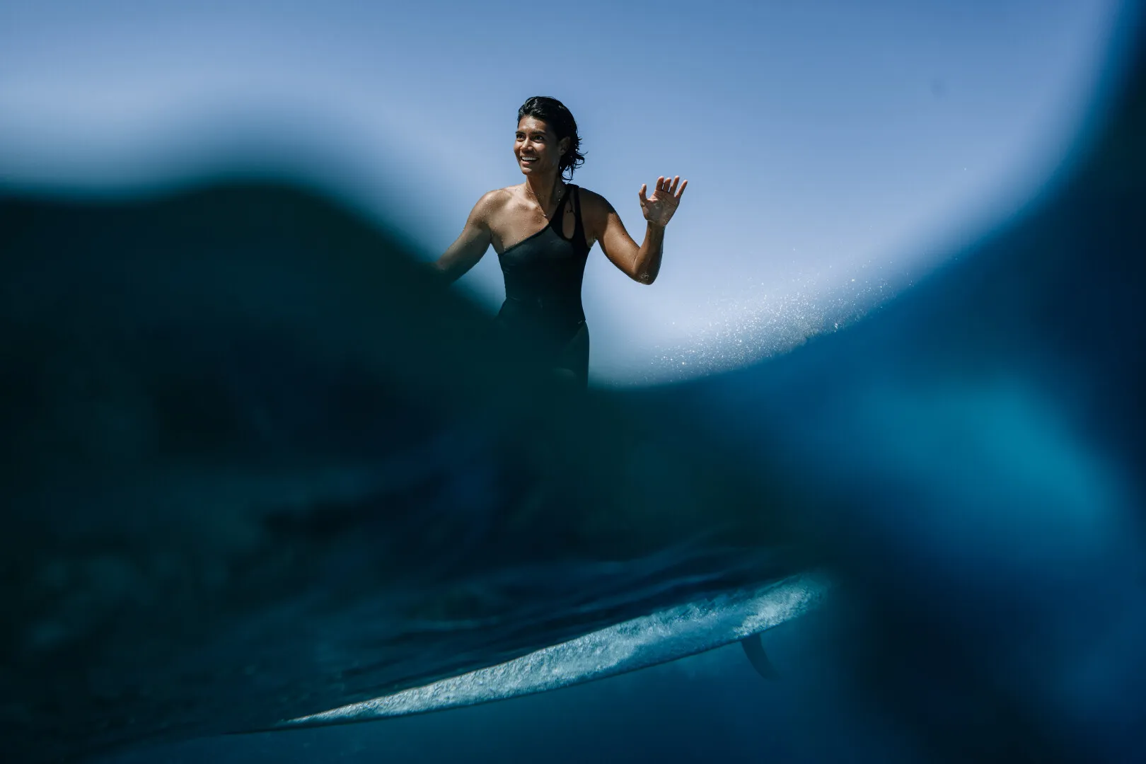 Smiling woman in a black swimsuit surfing on a blue wave with blurred water foreground.