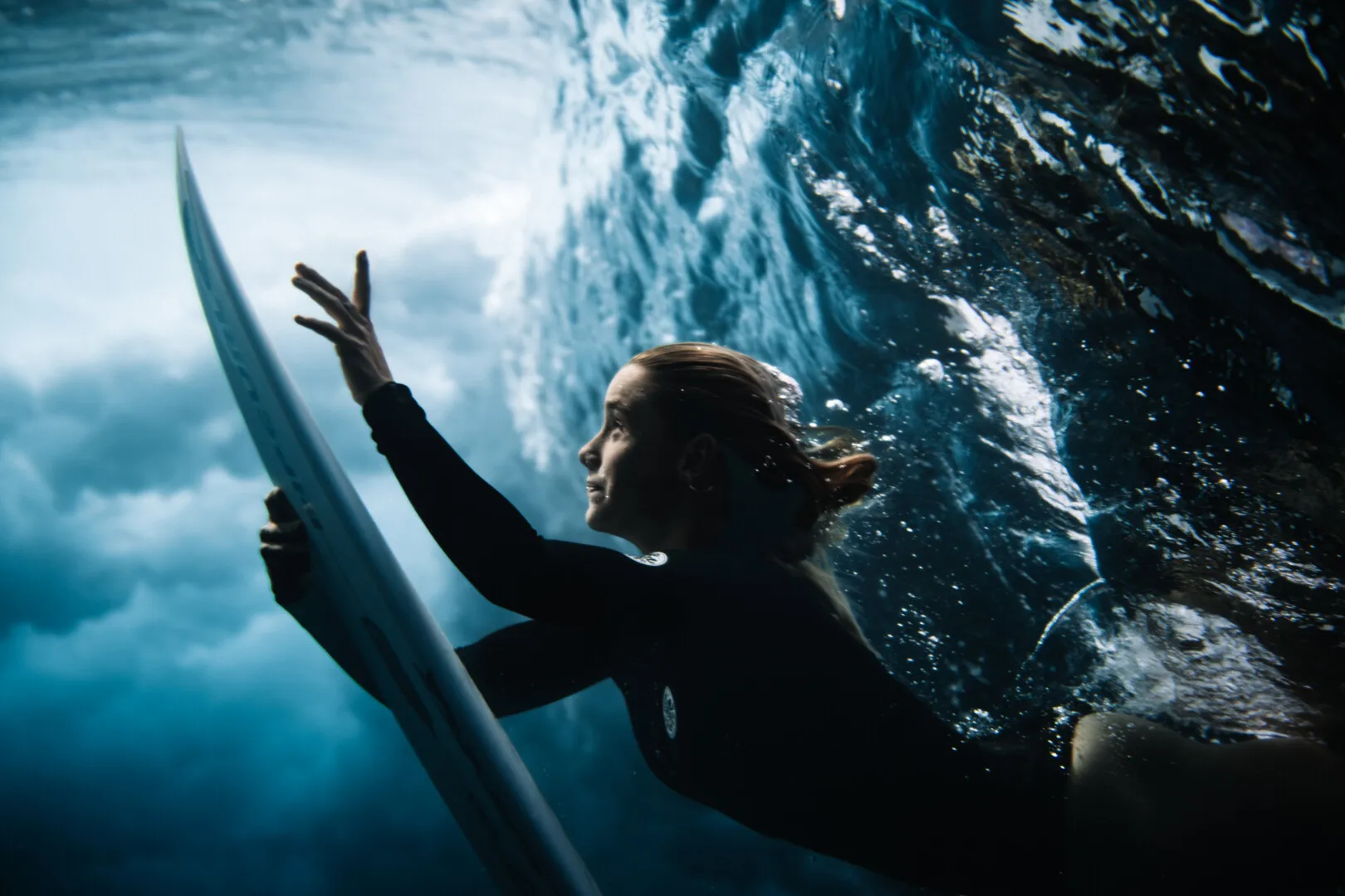 Female surfer underwater holding a surfboard, surrounded by bubbles and waves.