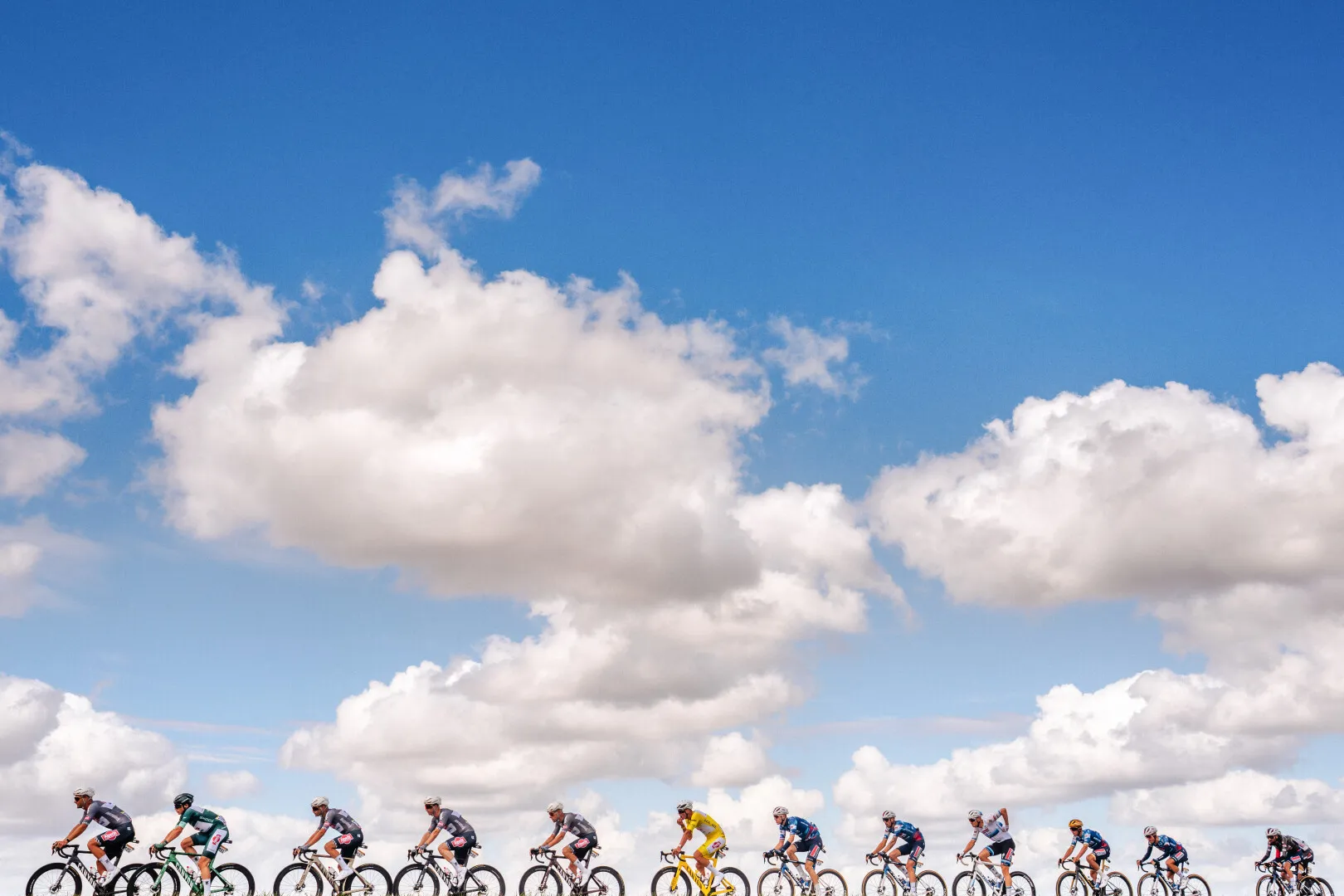 Group of professional cyclists riding in a line under a blue sky with scattered clouds.