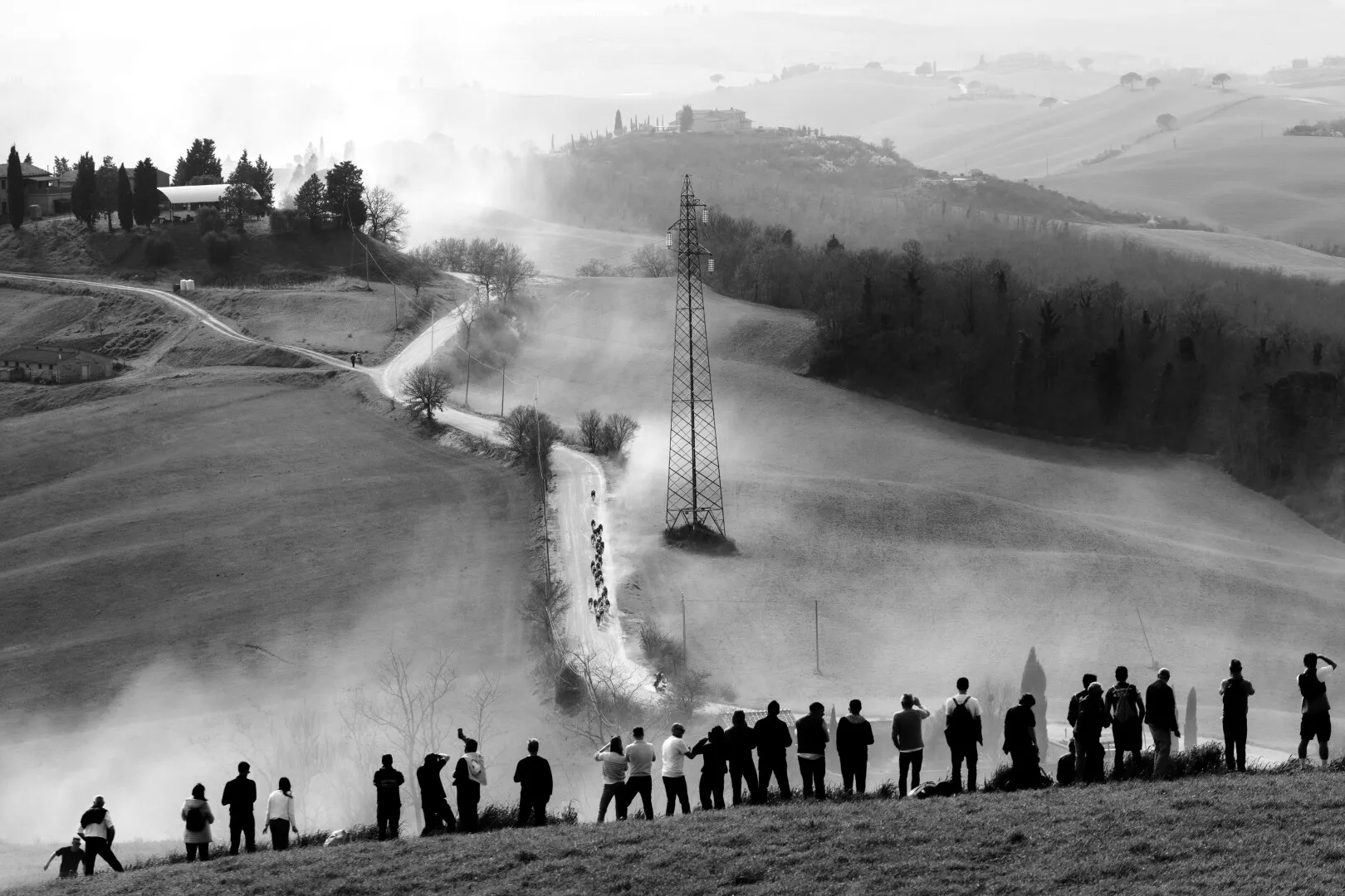 Silhouettes of people standing on a hill watching a dirt road winding through a foggy countryside with cyclists racing along it.