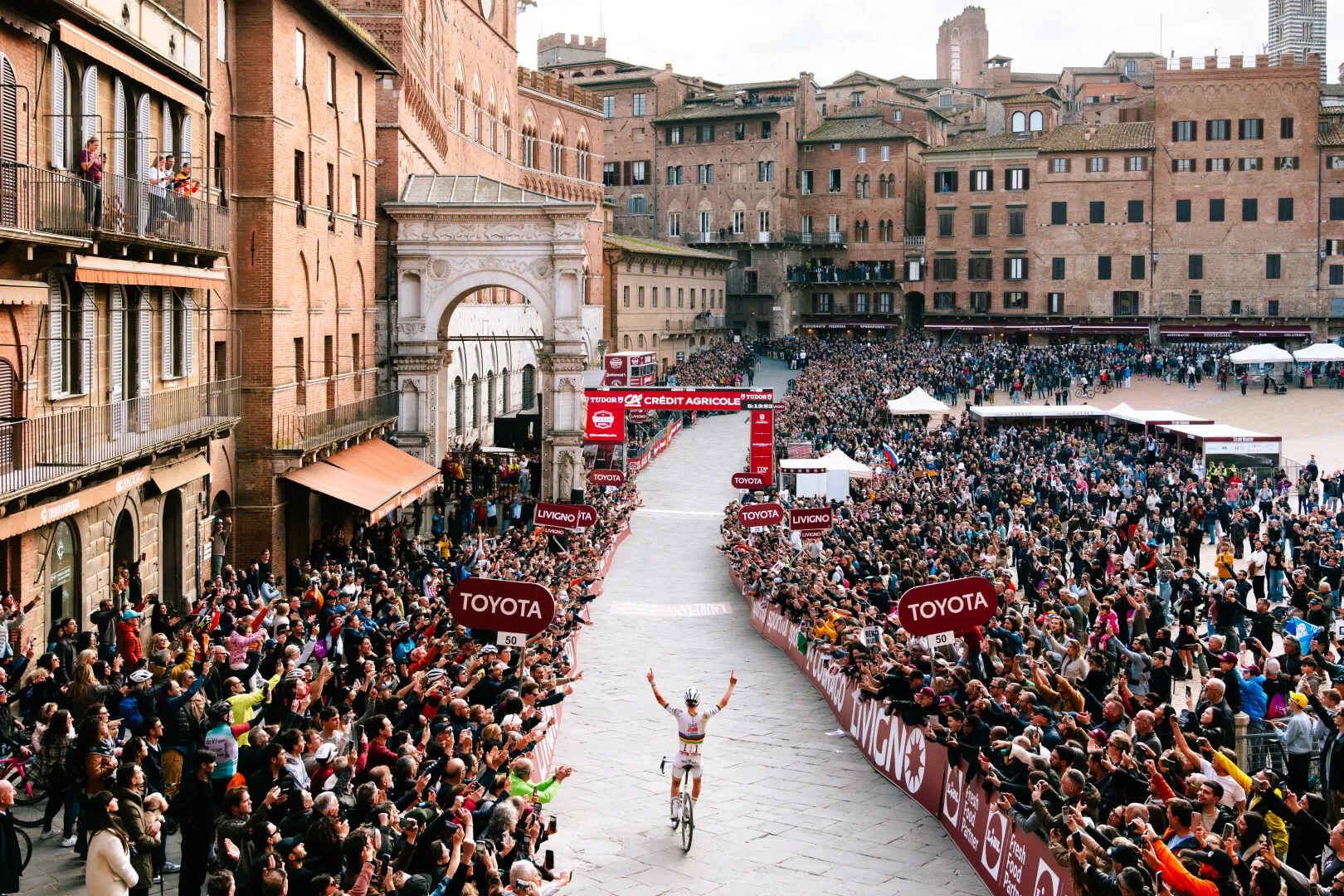 Cyclist crossing finish line in front of large cheering crowd in a historic city square surrounded by old brick buildings.