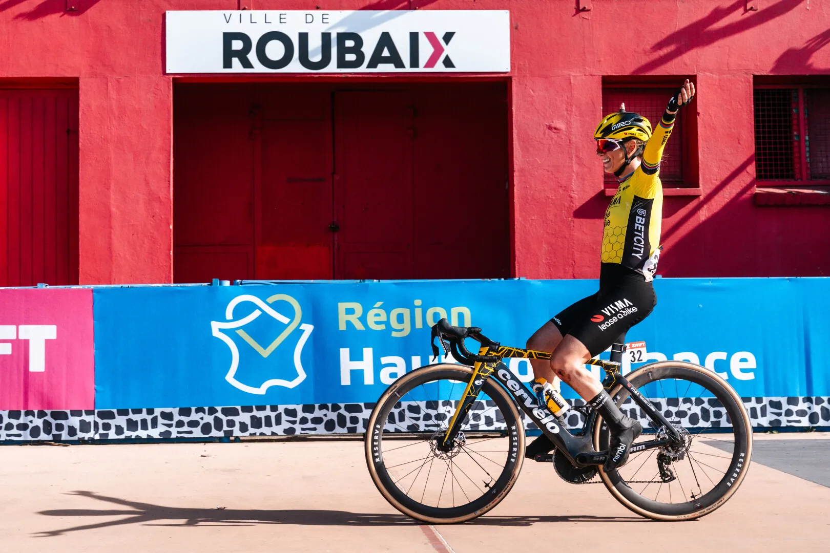 Cyclist in yellow and black jersey celebrating victory while riding a Cervelo bike in front of a red building with 'Ville de Roubaix' sign.